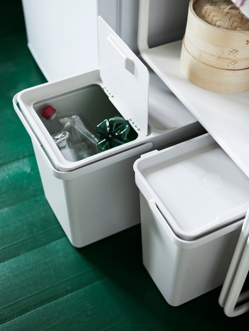 Two white HÅLLBAR kitchen bins under a shelf. One is pulled out with the lid open showing plastic waste ready for recycling.