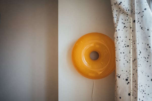 白い壁と水玉模様のカーテンの間に、丸いドーナツ形の黄色いウォールライトが取り付けられている。