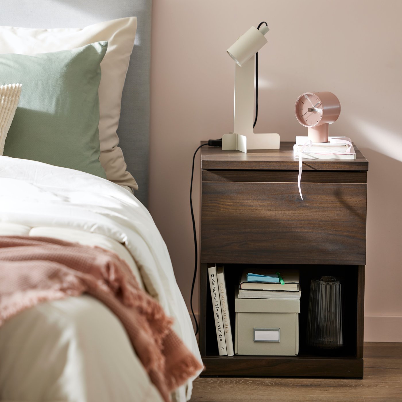 A dark brown oak effect STORKLINTA bedside table with a pale pink BONDTOLVAN alarm clock and beige FLOTTILJ desk lamp on top.