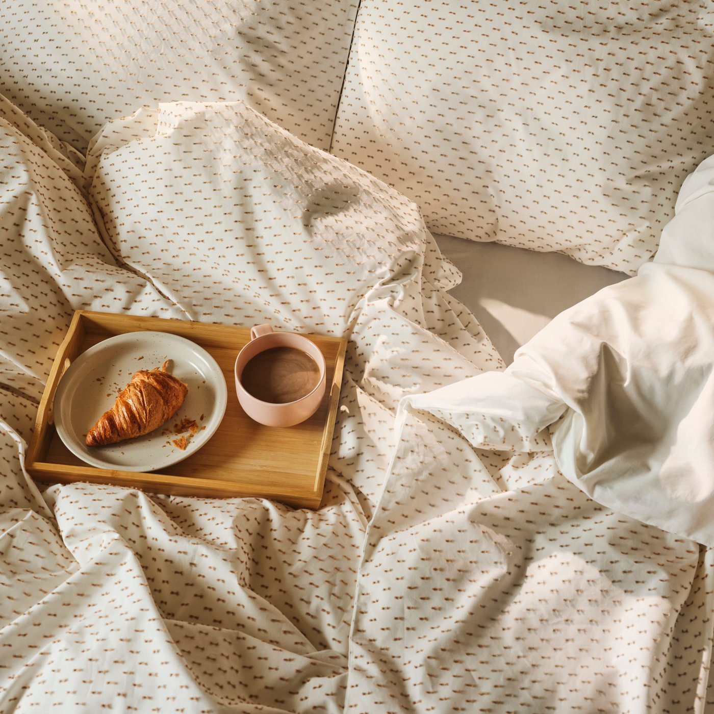 A bamboo FRUKTSKAL tray, with a cup of coffee and a croissant on a plate, sits on a bed with BANDKNIPPE bed linen.