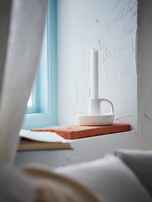 A white candlestick is placed on an orange surface next to a book, all of which set on a white windowsill.