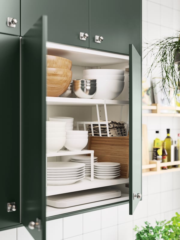 A wall cabinet with deep green kitchen fronts and doors ajar showing plates, bowls, a shelf insert and a clip-on basket.