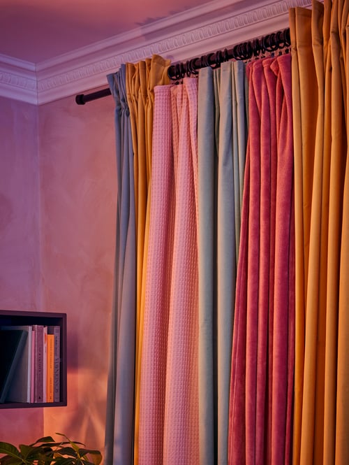 In einem Zimmer sind am Fenster abdunkelnde SANELA Gardinen zur Seite geschoben. Dazwischen hängt eine TRATTVIVA Tagesdecke in Dunkelrosa an Gardinenringen mit Klemme und Haken. 	
