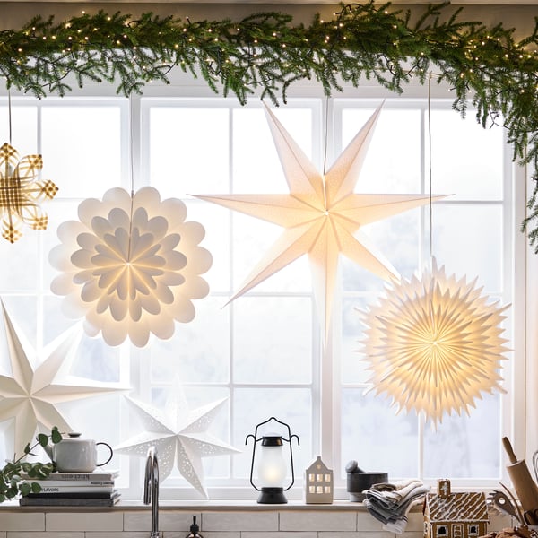 Una ventana con muchas lámparas en forma de estrella STRÅLA encendidas. Sobre la ventana hay una guirnalda en forma de planta con luces.