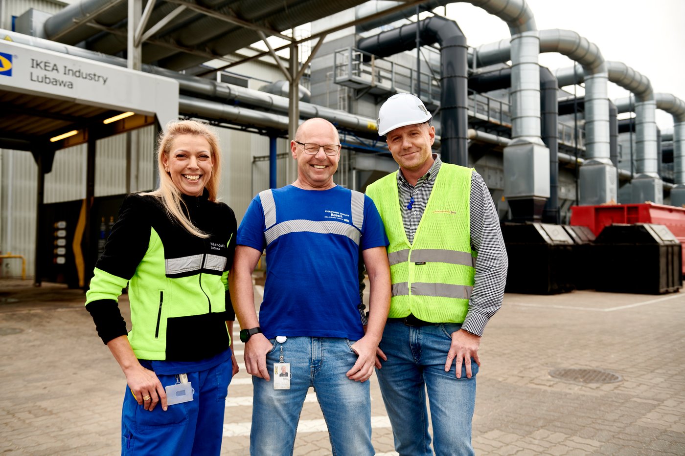 Trzech uśmiechających się pracowników - jedna kobieta i dwóch mężczyzn, pozujących na tle fabryki IKEA Industry Lubawa