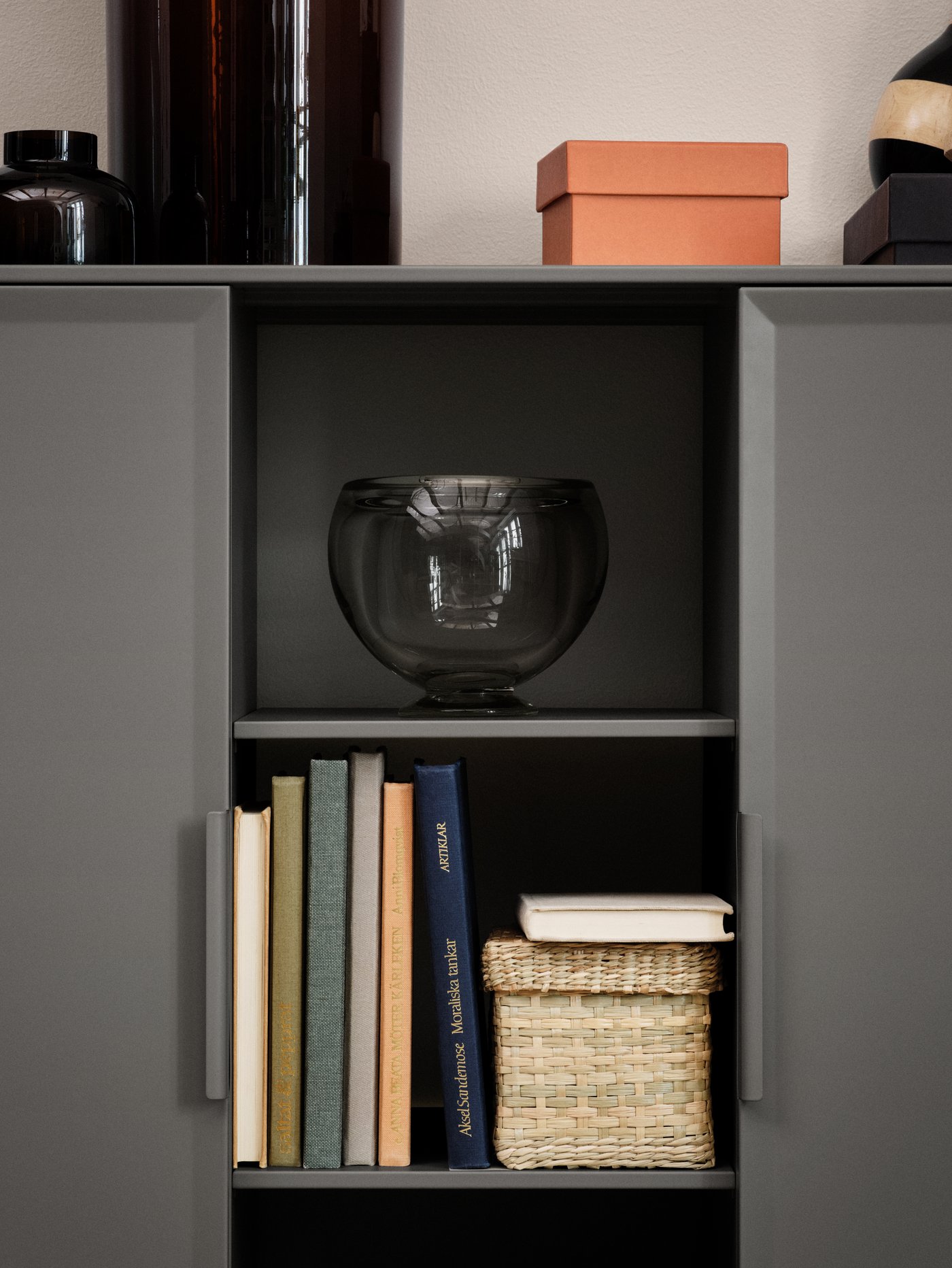 The open shelves of a grey TULLSTORP cabinet holding a vase, books and a basket. Boxes and glass vases are on top.