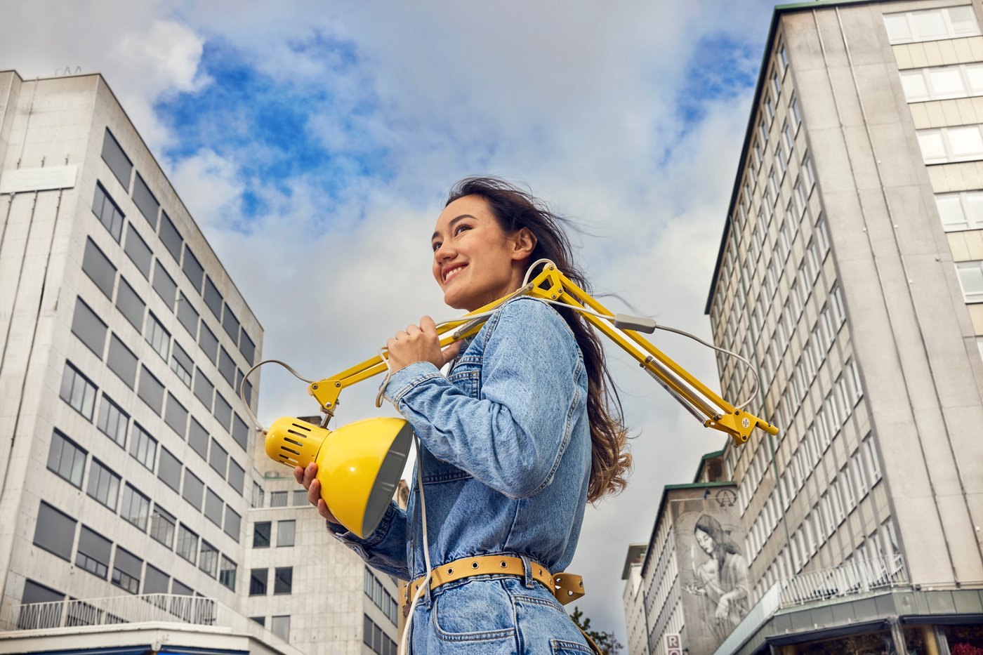 Mujer sonriente sosteniendo una lámpara amarilla sobre los hombros en una calle urbana entre edificios altos. Vista en contrapicado con cielo nublado.