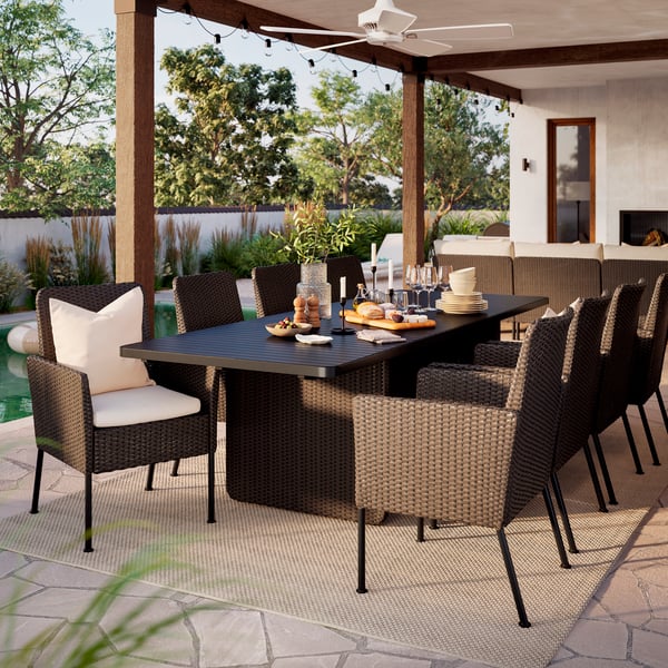 A large outdoor space with dark grey VITTSKÄR table and eight rattan dining chairs under a roof. Sun is shining.
