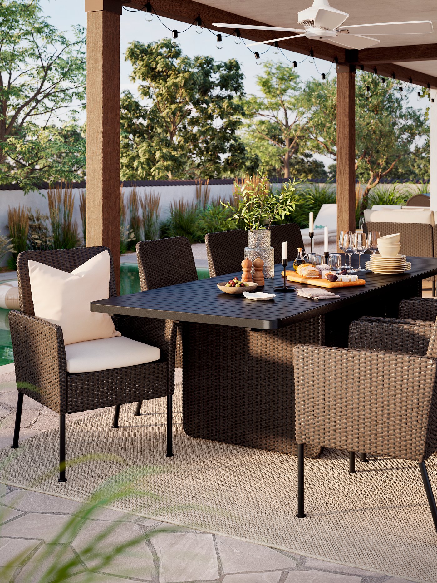 En stor utomhusyta med ett mörkgrått VITTSKÄR-bord och åtta matstolar i rotting under ett tak. Solen skiner.