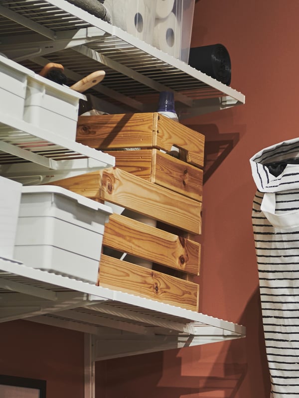 Different sizes of KNAGGLIG boxes and SOCKERBIT boxes with lids stand on white shelves which are attached to a red wall.
