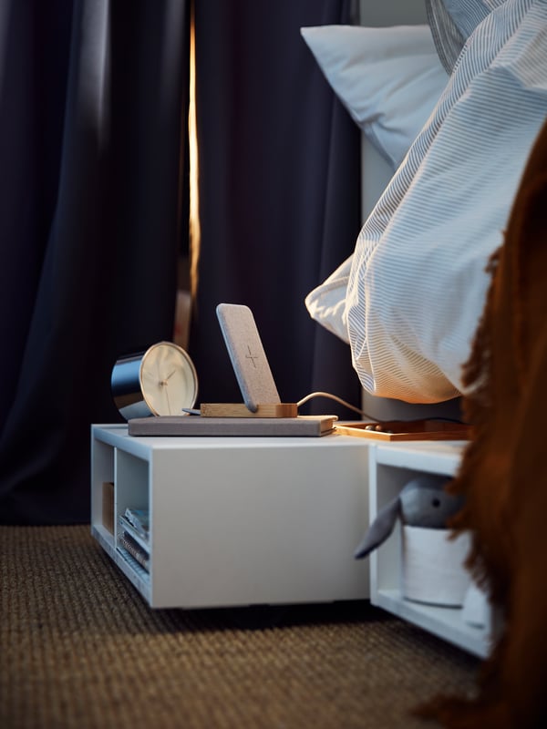 Two white FREDVANG underbed storage units/bedside tables holding books and various other items, on a jute rug under a bed.
