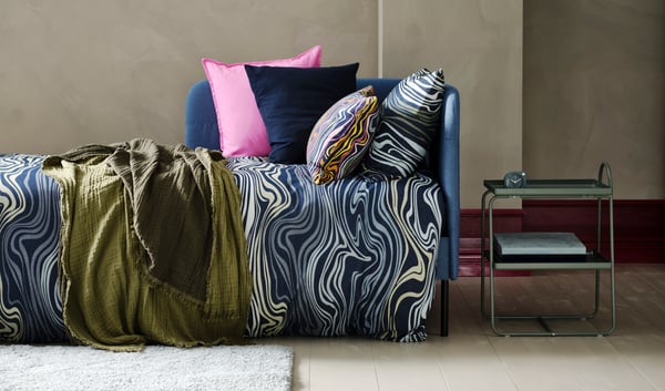 Estructura de cama con ropa de cama azul y blanca con estampado de ondas, cojines de colores azules y rosas y una mesa auxiliar en tonos verde, modelo HATTÅSEN.