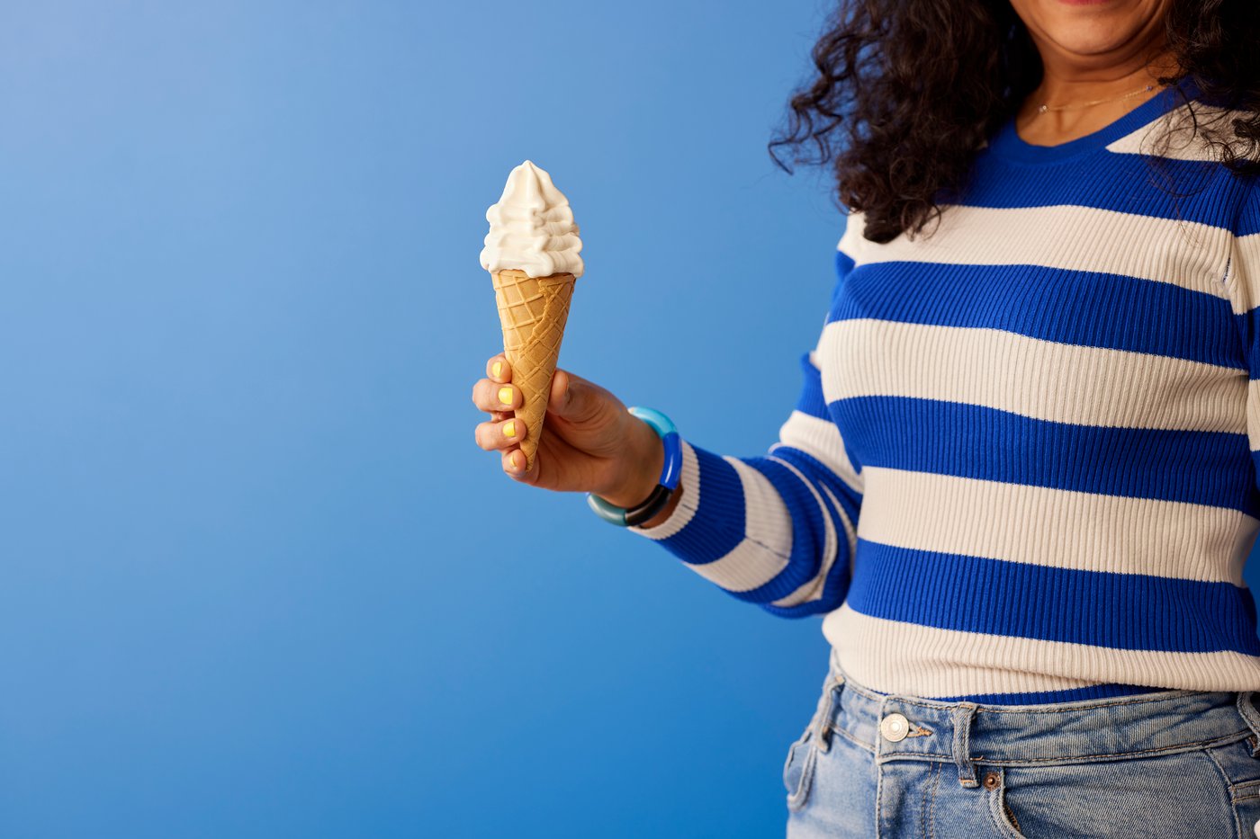 Frau in blau-weiß gestreiftem Pullover und blauer Jeans hält ein Softeis mit Vanillegeschmack vor einem einfarbig blauen Hintergrund.
