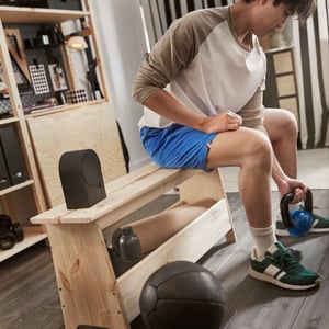 Osoba siedzi na sosnowej ławce i ćwiczy, podnosząc ciężary. Tuż obok, na ławce stoi czarny głośnik Blueooth NATTBAD.