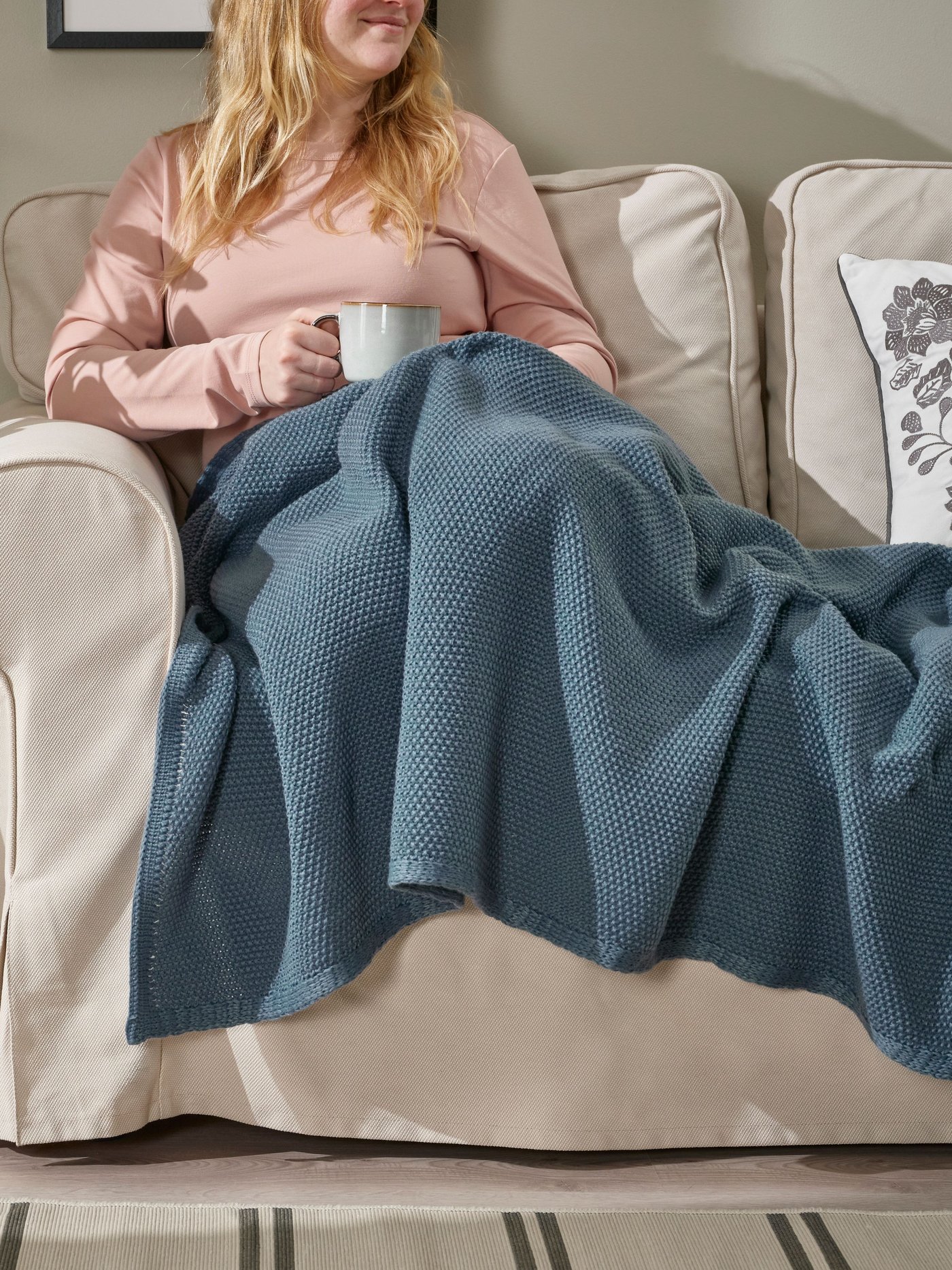 Eine Frau sitzt mit einer Tasse in der Hand auf einem Sofa und ist mit einer blaugrauer HUMLEMOTT Decke zugedeckt.