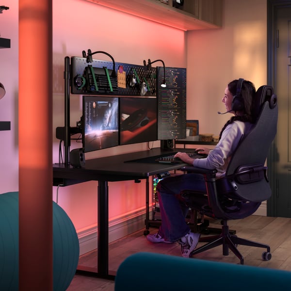 A person in a BÄSTBOLL gaming chair is at a MÅLOMRÅDE gaming desk. NÄVLINGE clamp spotlights are attached to the desk.