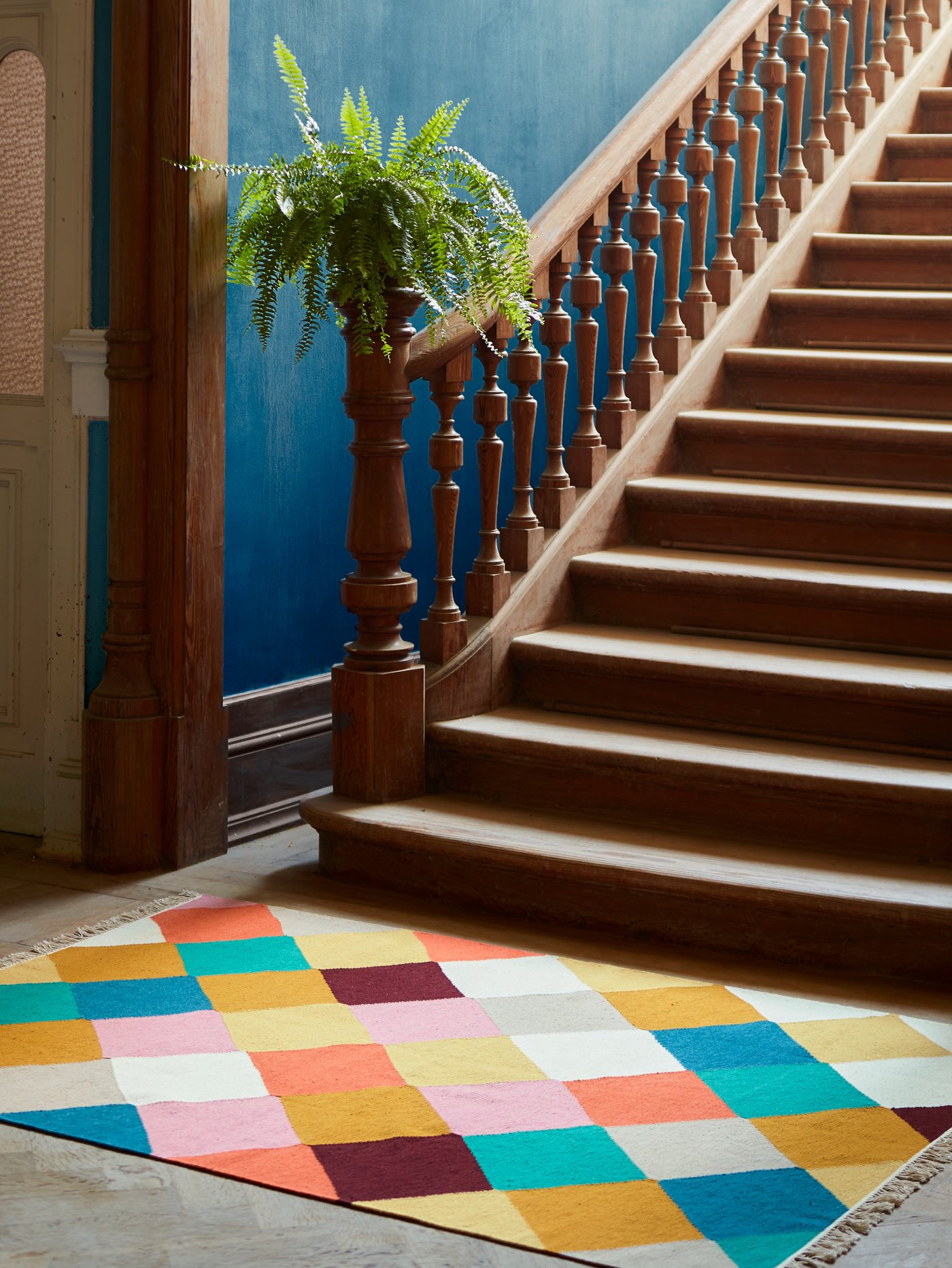 The handwoven VINDERÖD rug in wool made in a colourful, chequered pattern, placed at the bottom of a staircase.
