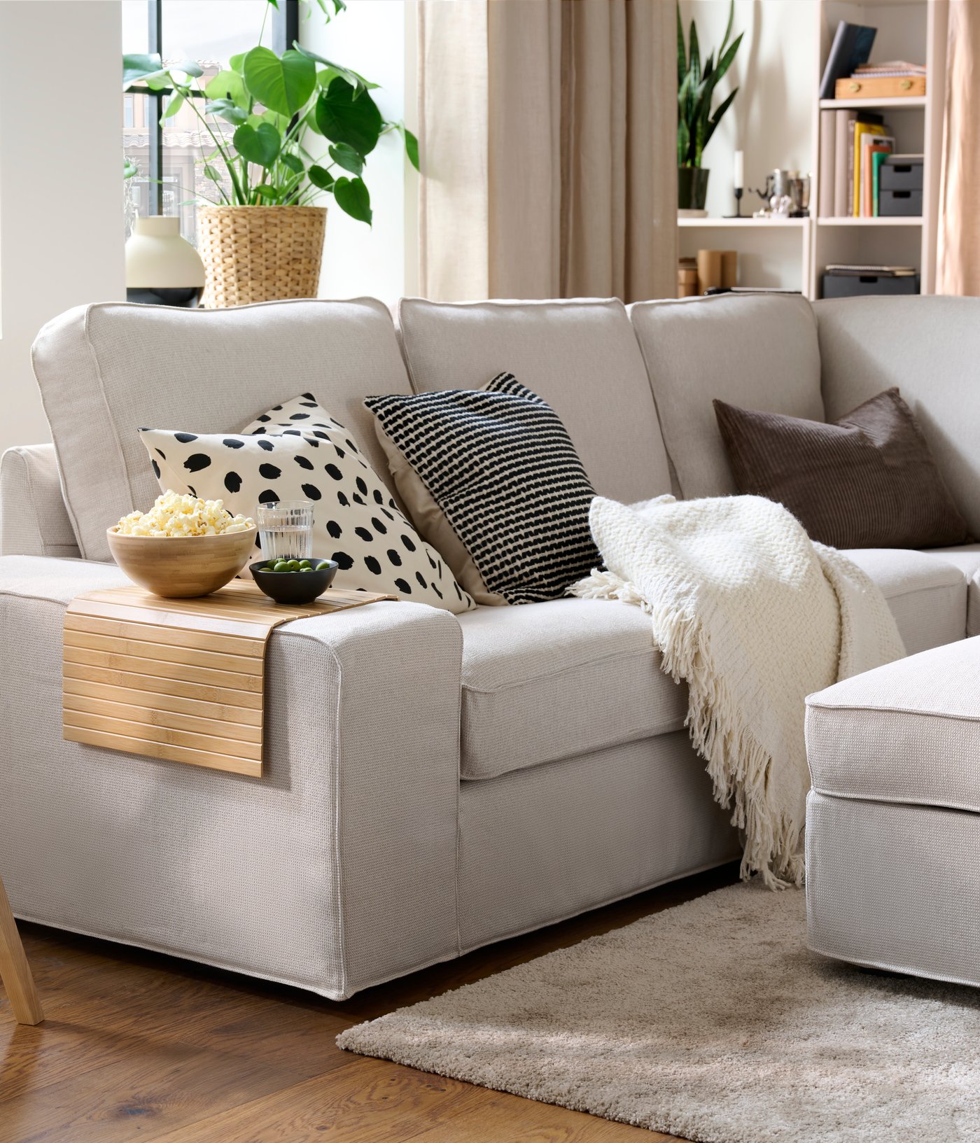 A light living room with a KIVIK corner sofa with colourful cushions, a throw, and a RÖDEBY armrest tray holding snack bowls.