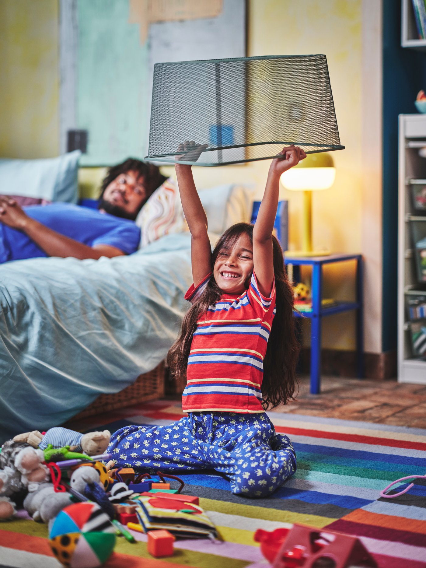A child on a floor next to a bed is holding a TROFAST bin upside down above her head. There are toys all over the floor.