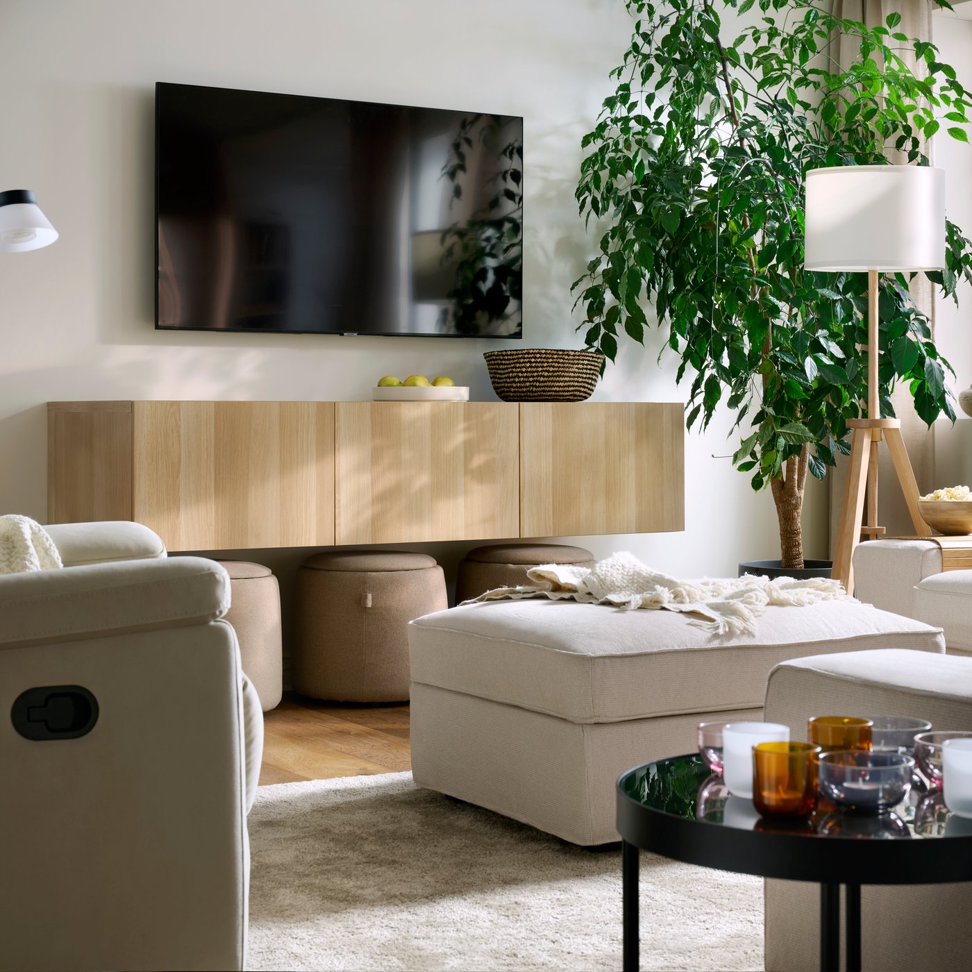 Ein leichtes, helles Wohnzimmer mit einem KIVK-Pouffe, einem SKULNÄS-Sessel, einem STOENSE-Teppich, einem an der Wand montierten Fernseher und einer großen, blattreichen Pflanze.