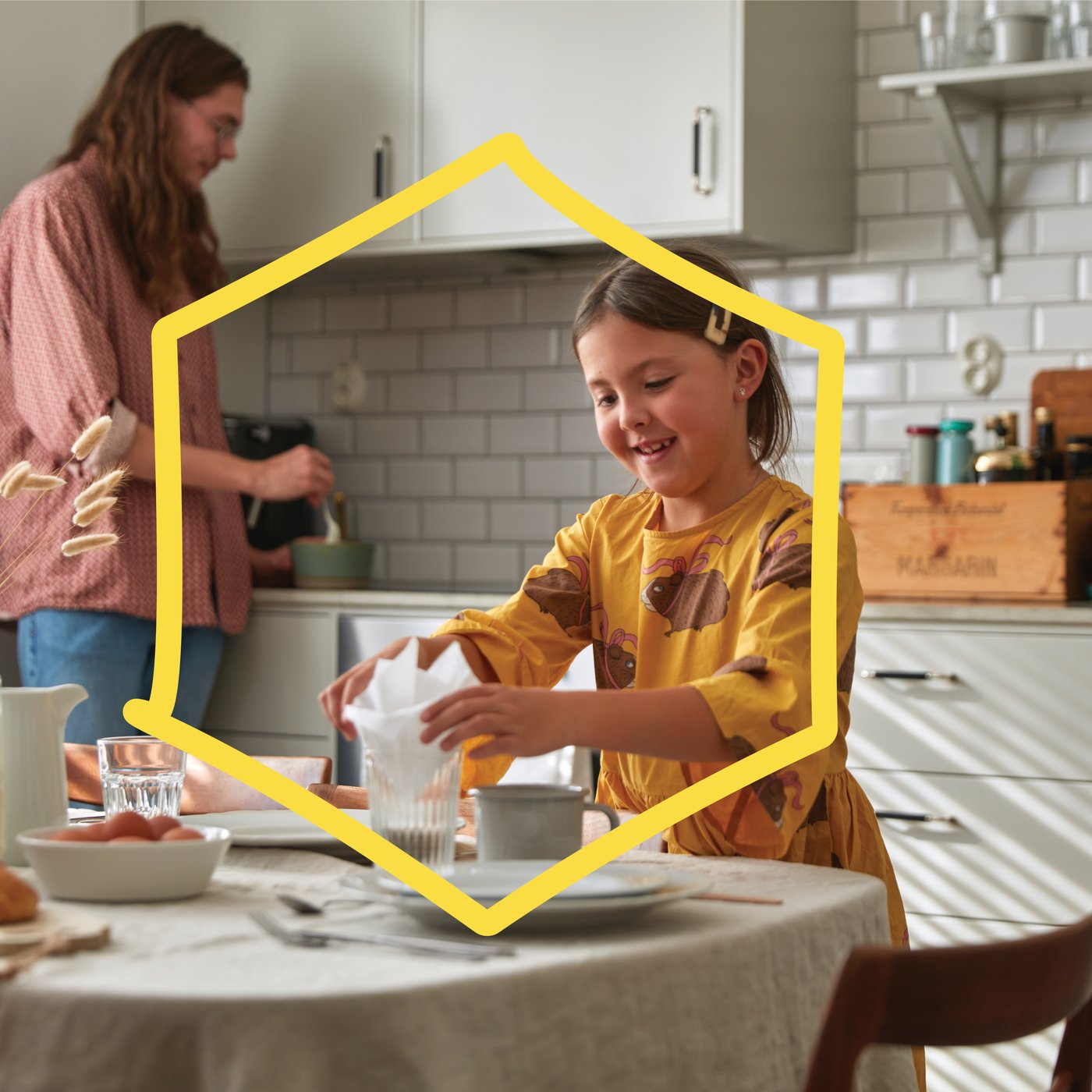 A child in a yellow dress places a napkin into a glass at a table, while another person stirs ingredients in a bowl in a bright kitchen with white subway tiles.