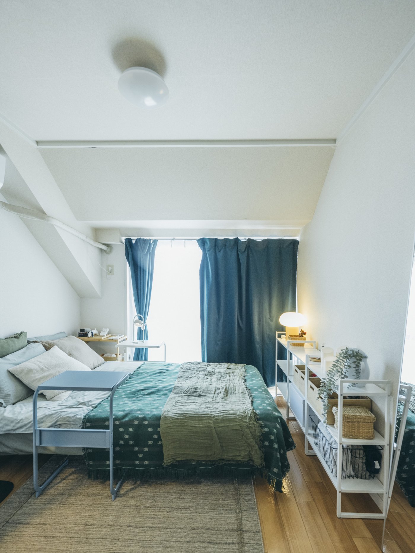 緑の寝具がかけられたベッドが置かれ、青いカーテンがかかった窓から日が差している。