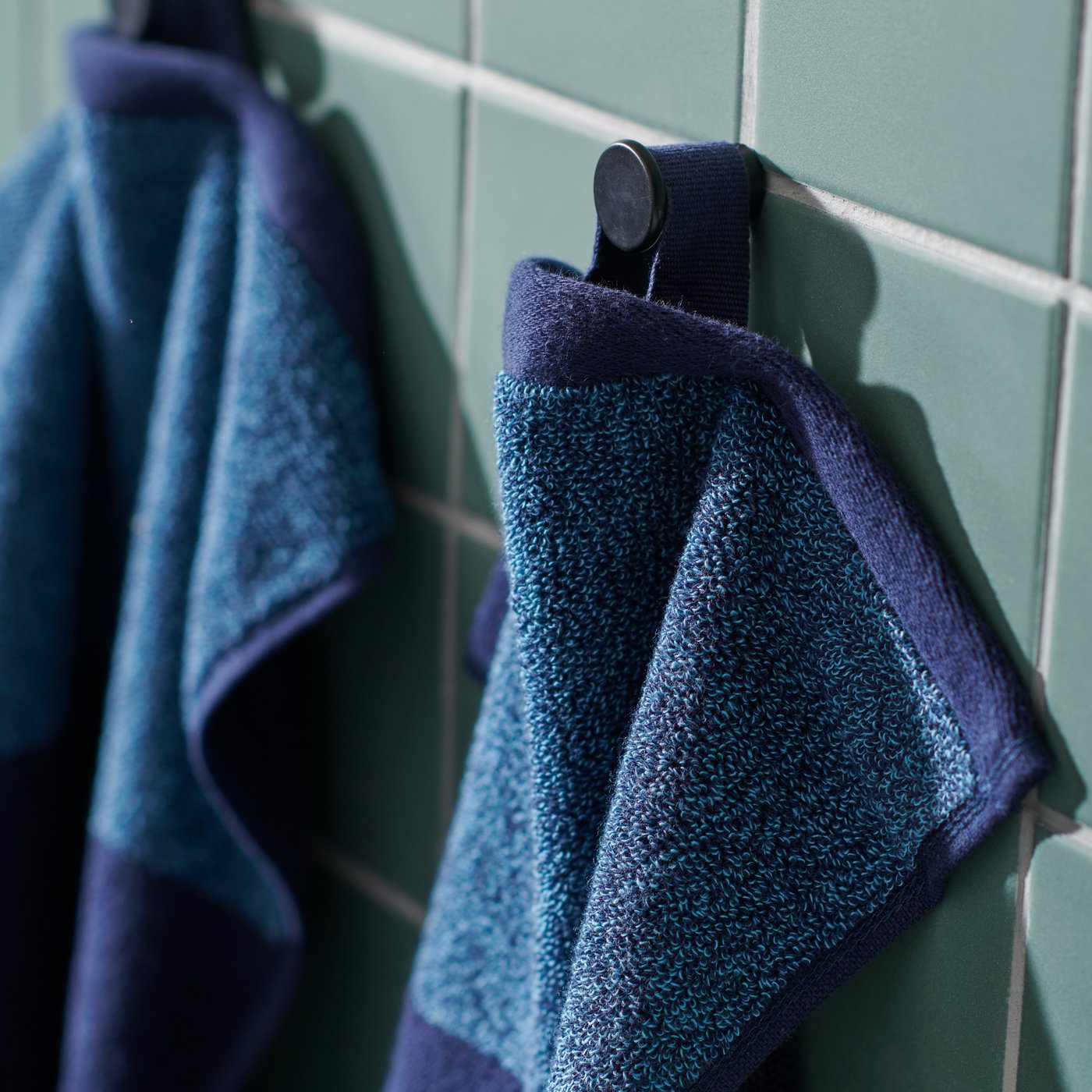 Dua tuala HIMLEÅN berwarna biru-mélange tergantung di tombol gelap pada dinding bilik mandi jubin berwarna hijau.