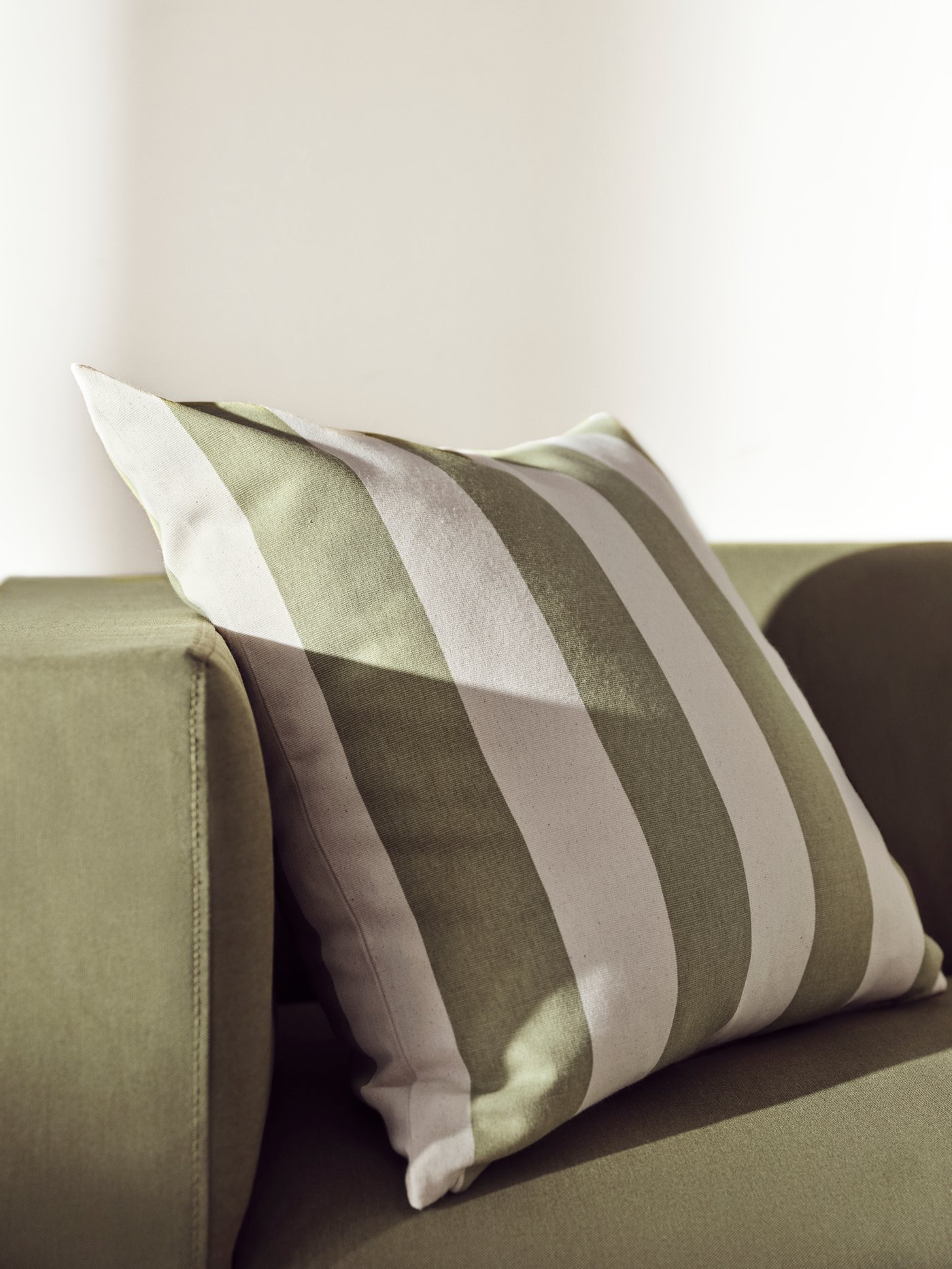 Un coussin HILDAMARIA à rayures vertes et blanches est placé sur un canapé vert sous un rayon de soleil.