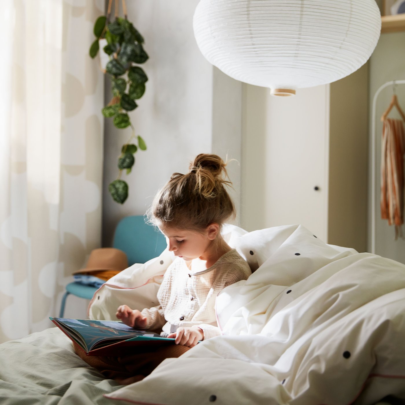 A girl reading on a bed with a quilt around her, with a dotted white/pink VÄNKRETS quilt cover, and two white pendants above.