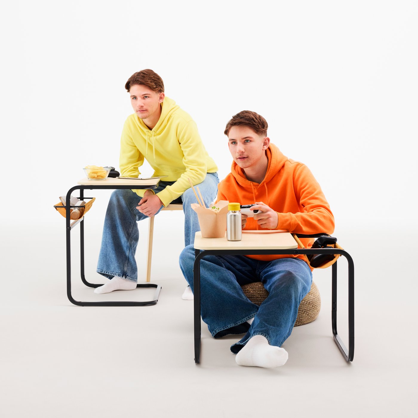 Two teenage twins, each sitting at an OLSERÖD side table, one in tall side table mode, the other in low coffee table mode.