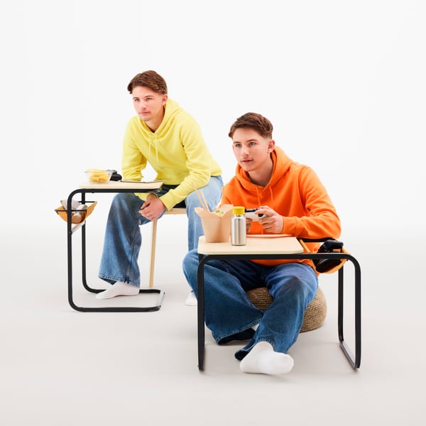 Two teenage twins, each sitting at an OLSERÖD side table, one in tall side table mode, the other in low coffee table mode.