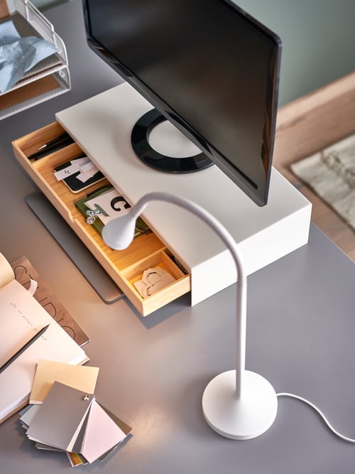 Un spațiu de lucru modern cu un suport pentru monitor bej cu sertar din oțel și bambus, deasupra unui birou dreptunghiular.