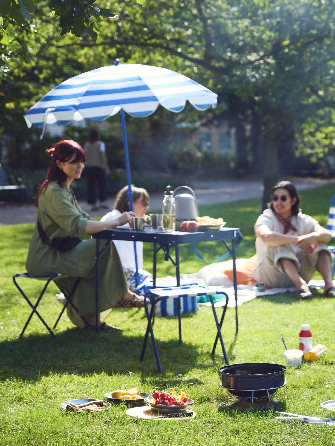 Friends enjoy a relaxing picnic and barbecue in the park, comfortably set up with STRANDÖN chairs, table, and parasol.