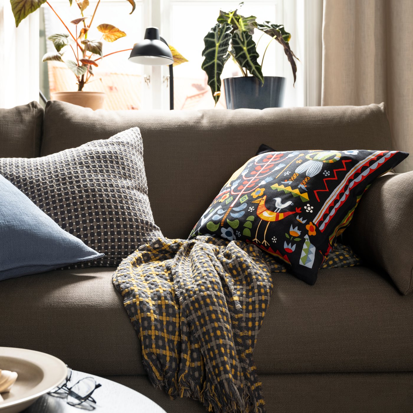 En HYLTARP sofa med en plaid i ternet mønster og nogle ekstra puder, en af dem med et flerfarvet TRATTKAKTUS pudebetræk.