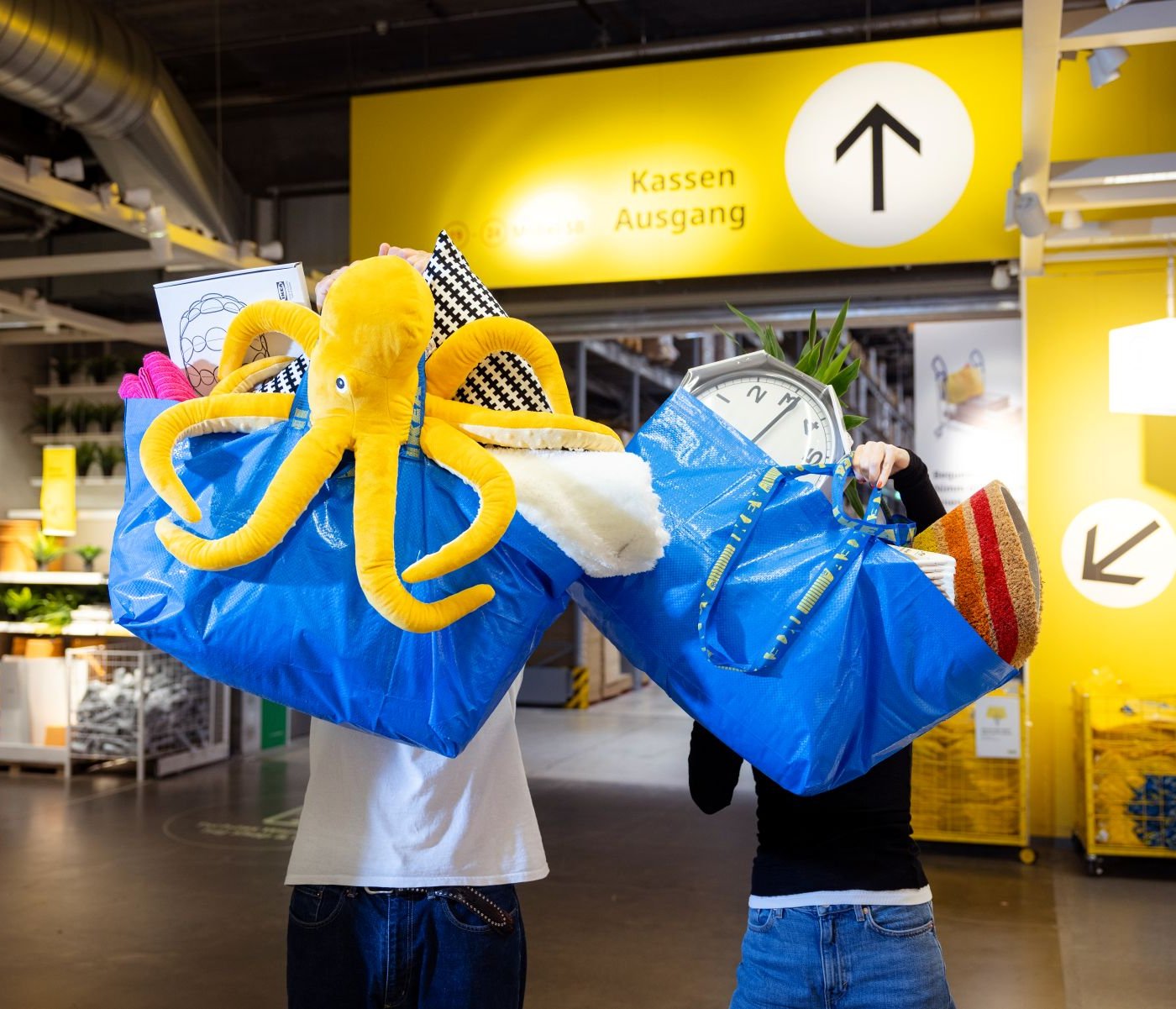 Zwei Personen tragen in einem IKEA Einrichtungshaus große blaue Einkaufstaschen voller Produkte, darunter ein auffälliger gelber Plüschtintenfisch, unter einem gelben Wegweiser mit der Aufschrift „Kassen Ausgang“.