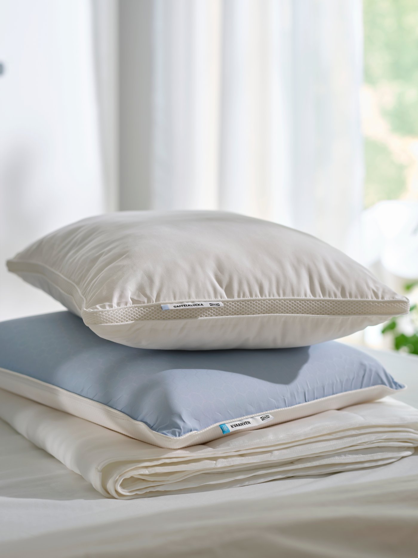 Ein GAFFELKLOCKA Kissen und ein KVARNVEN ergonomisches Kissen sind auf einer Bettdecke auf einem Bett gestapelt.
