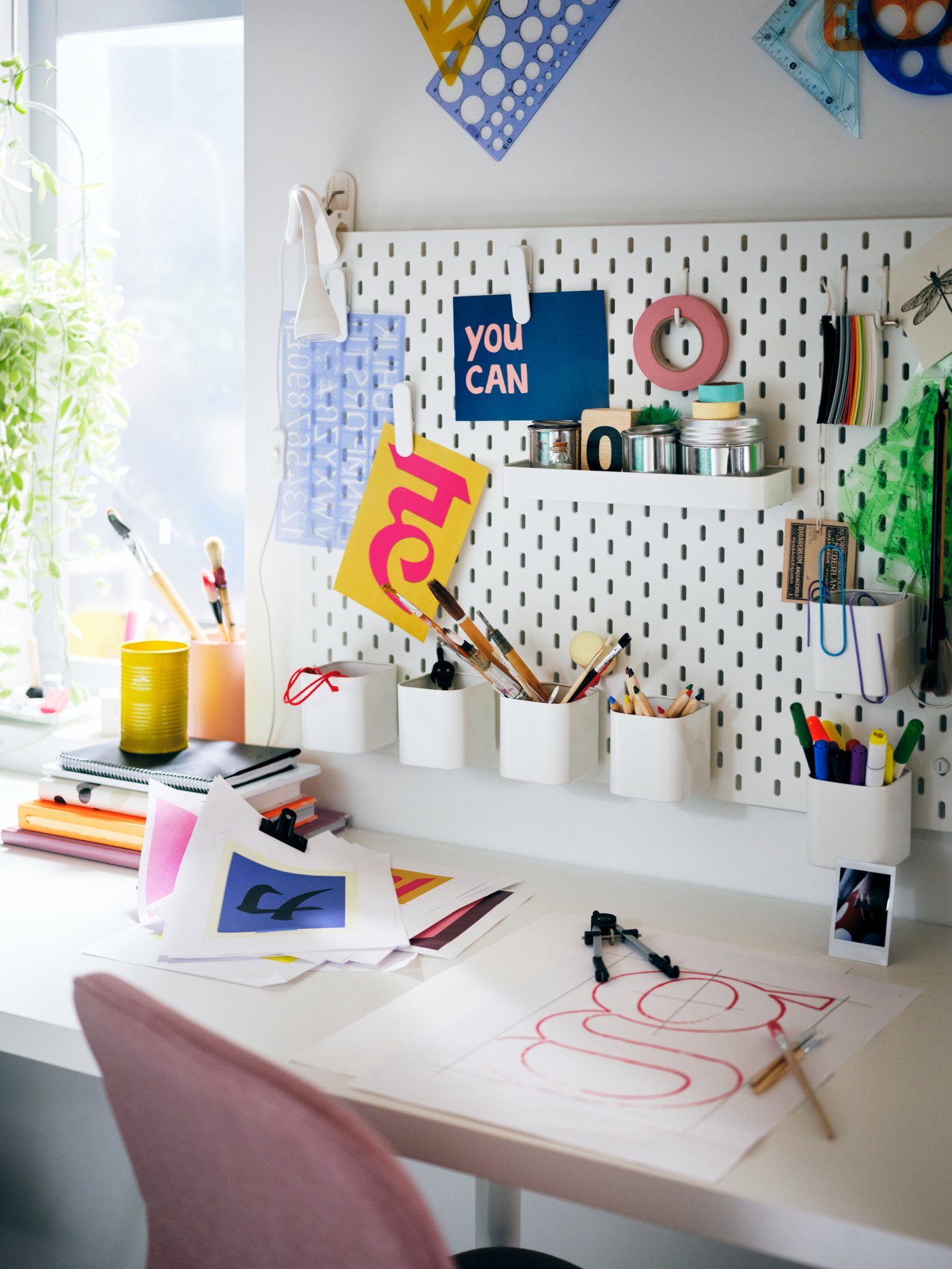 A white SKÅDIS pegboard filled with colourful accessories, over a white LAGKAPTEN tabletop and pink LÅNGFJÄLL swivel chair.