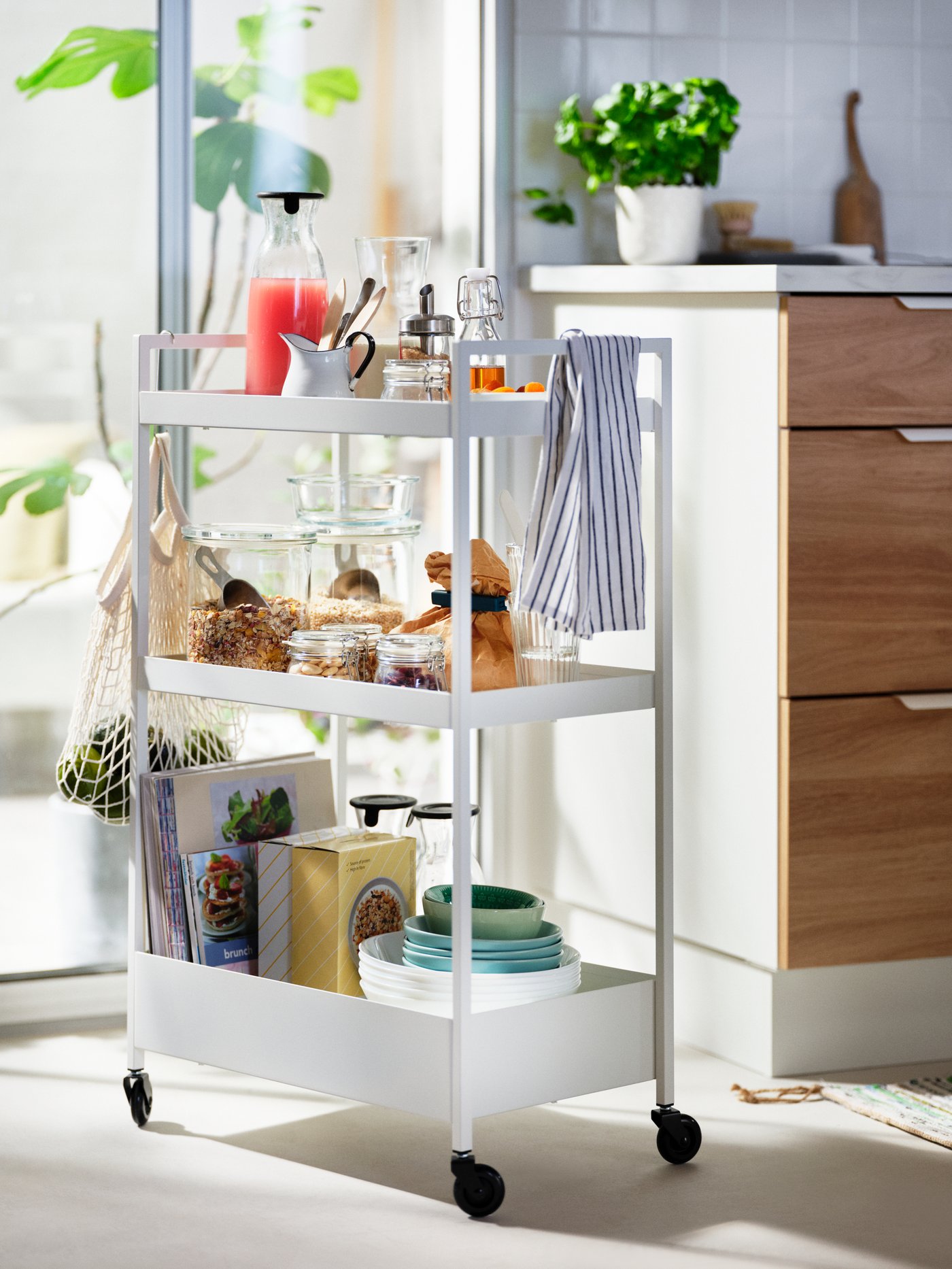A white, sunlit NISSAFORS trolley loaded with food and accessories standing by a kitchen worktop and next to a glass door.