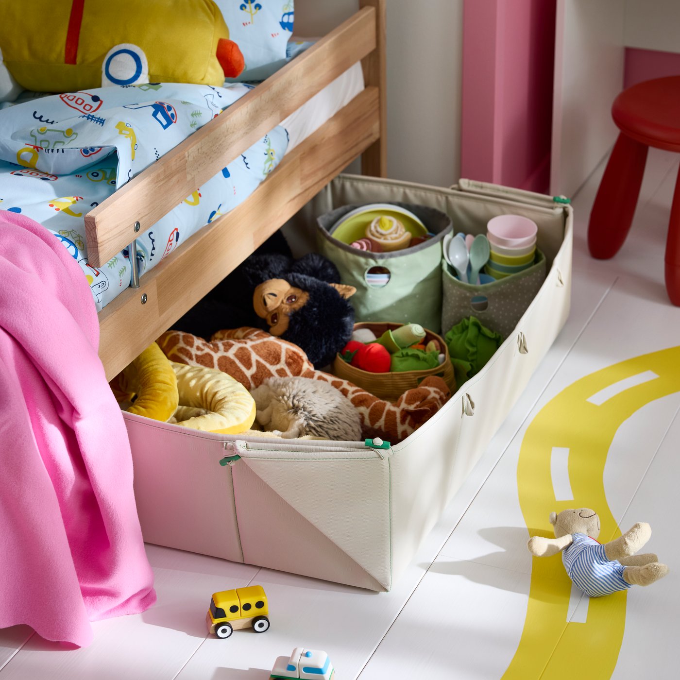 Under a bed sits a GREJSIMOJS mat and storage box filled with soft toys in a children’s room.