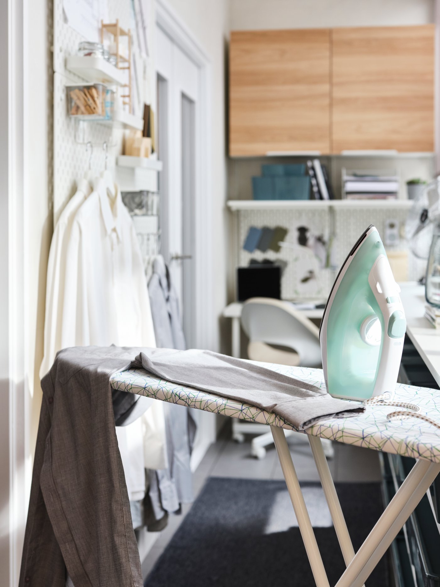 Una plancha y una camisa gris en una tabla de planchar RUTER en blanco, camisas colgando en almacenaje de pared blanco y armarios ENHET detrás.