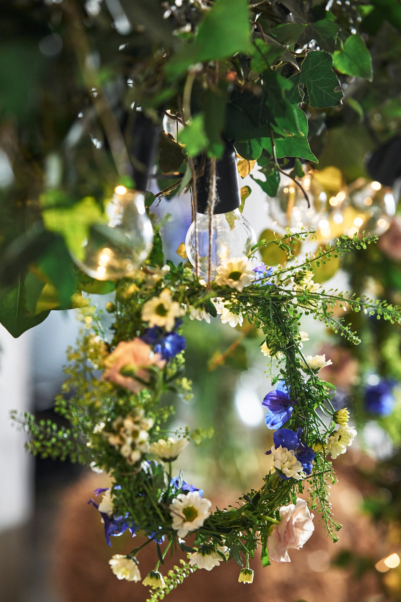 Des ampoules suspendues sous des feuilles vertes, entourées d'une couronne décorative composée de fleurs blanches, jaunes et bleues ainsi que de feuillage délicat, baignées d'une lumière chaude et naturelle à l'extérieur.