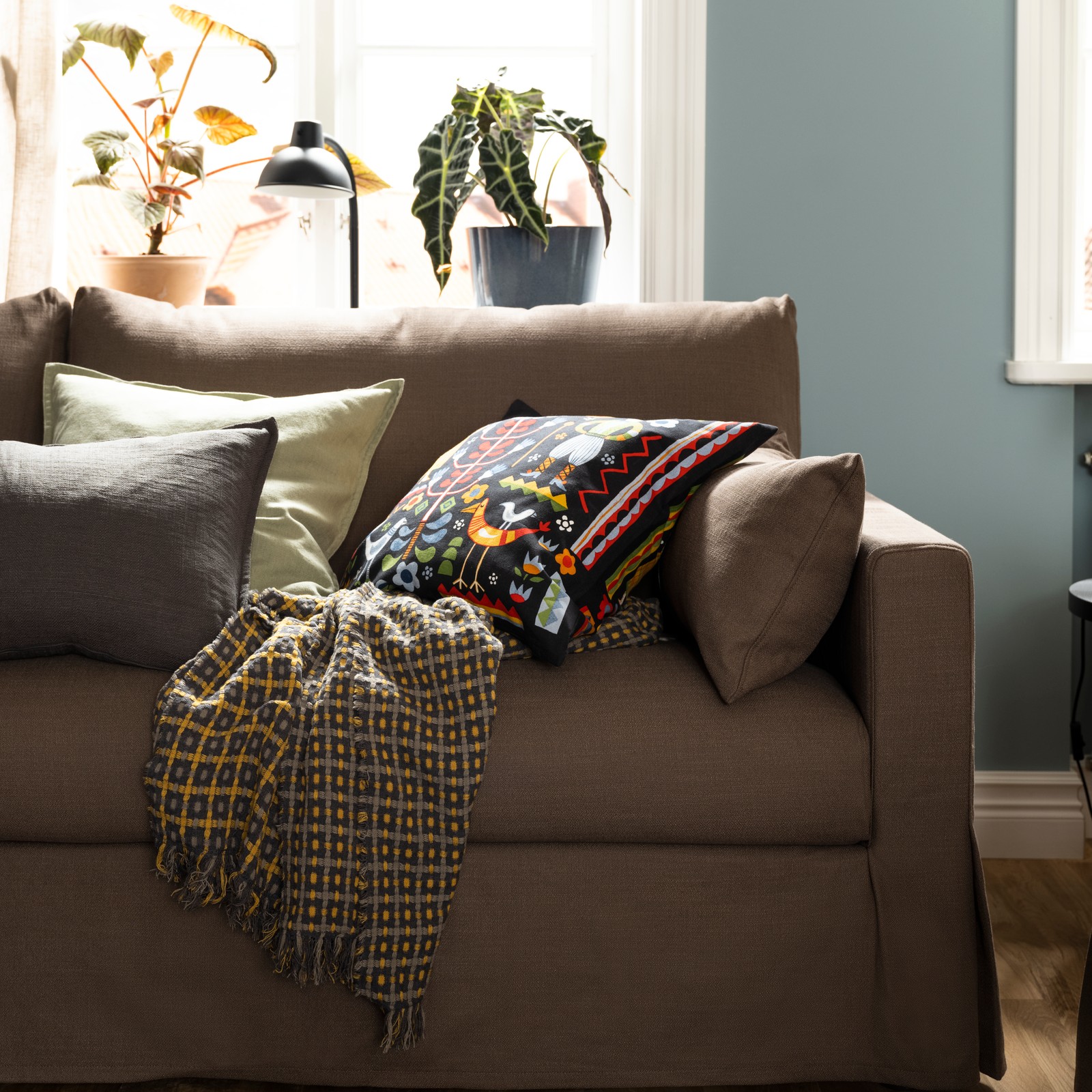 A FLIKBRUNÖRT throw and some cushions, including one with a multicolour TRATTKAKTUS cushion cover, lie on a HYLTARP sofa.