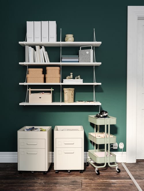 A green wall has four rows of white BERGSHULT/PERSHULT wall shelves mounted on it, above three storage units on castors.