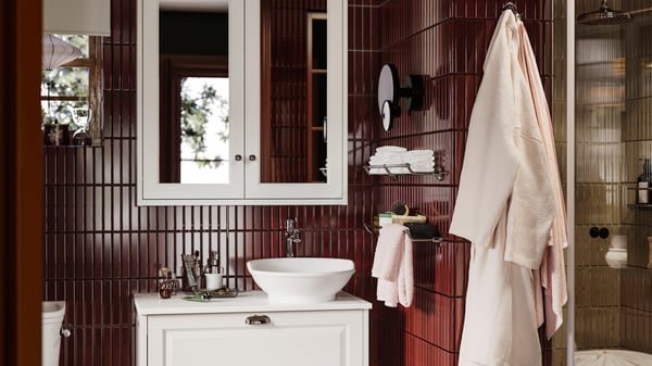A bathroom featuring TÄNNFORSEN mirror cabinet with drawers, folded towels, and hanging bathrobe on maroon tiled walls.