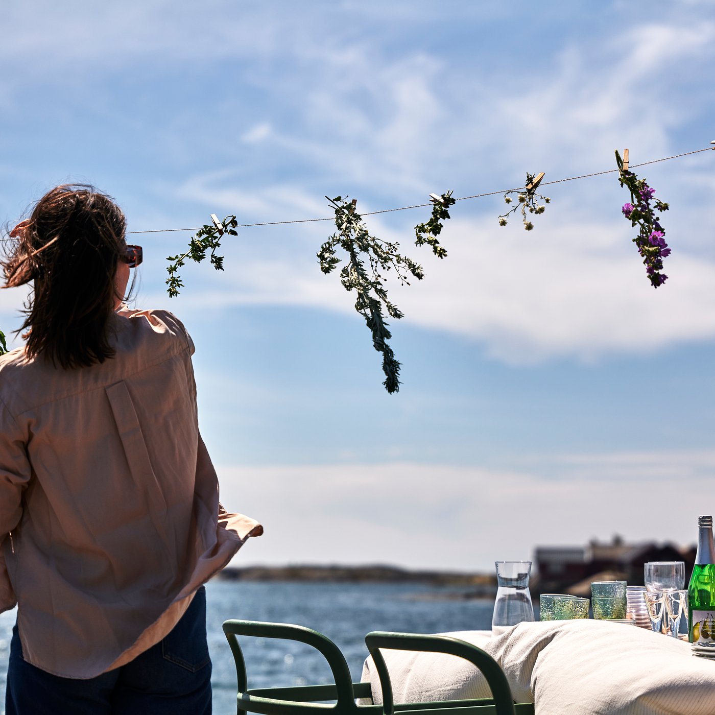Una persona penja flors en un fil a l'aire lliure, amb una taula parada al costat d'un cos d'aigua en un dia assolellat.