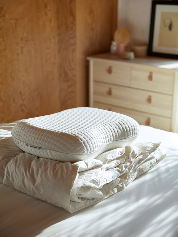 A white ergonomic pillow on top of a pile of white bedlinen on a white bed with daylight coming in from the window.