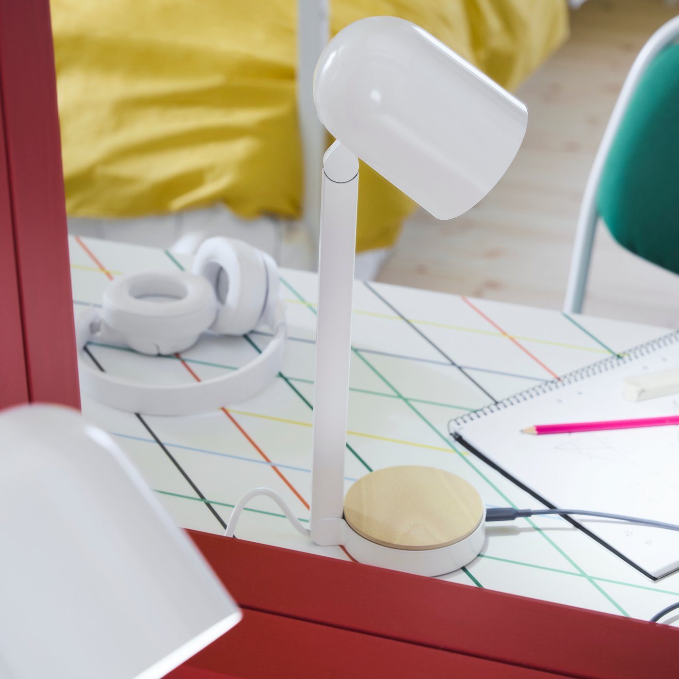 The white ENSMÄRKE desk lamp placed on a desk with a graphic pattern with a bed in the background made with yellow bedlinen.