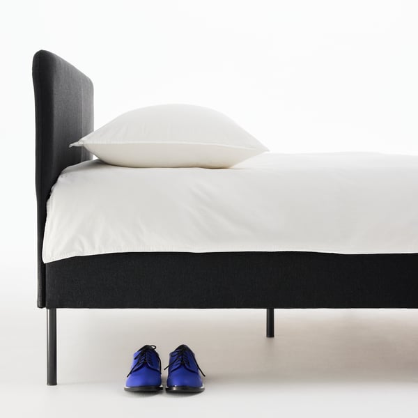 Close-up of a black SLATTUM bed, neatly made with white bed linen. A pair of blue dress shoes is underneath the bed.