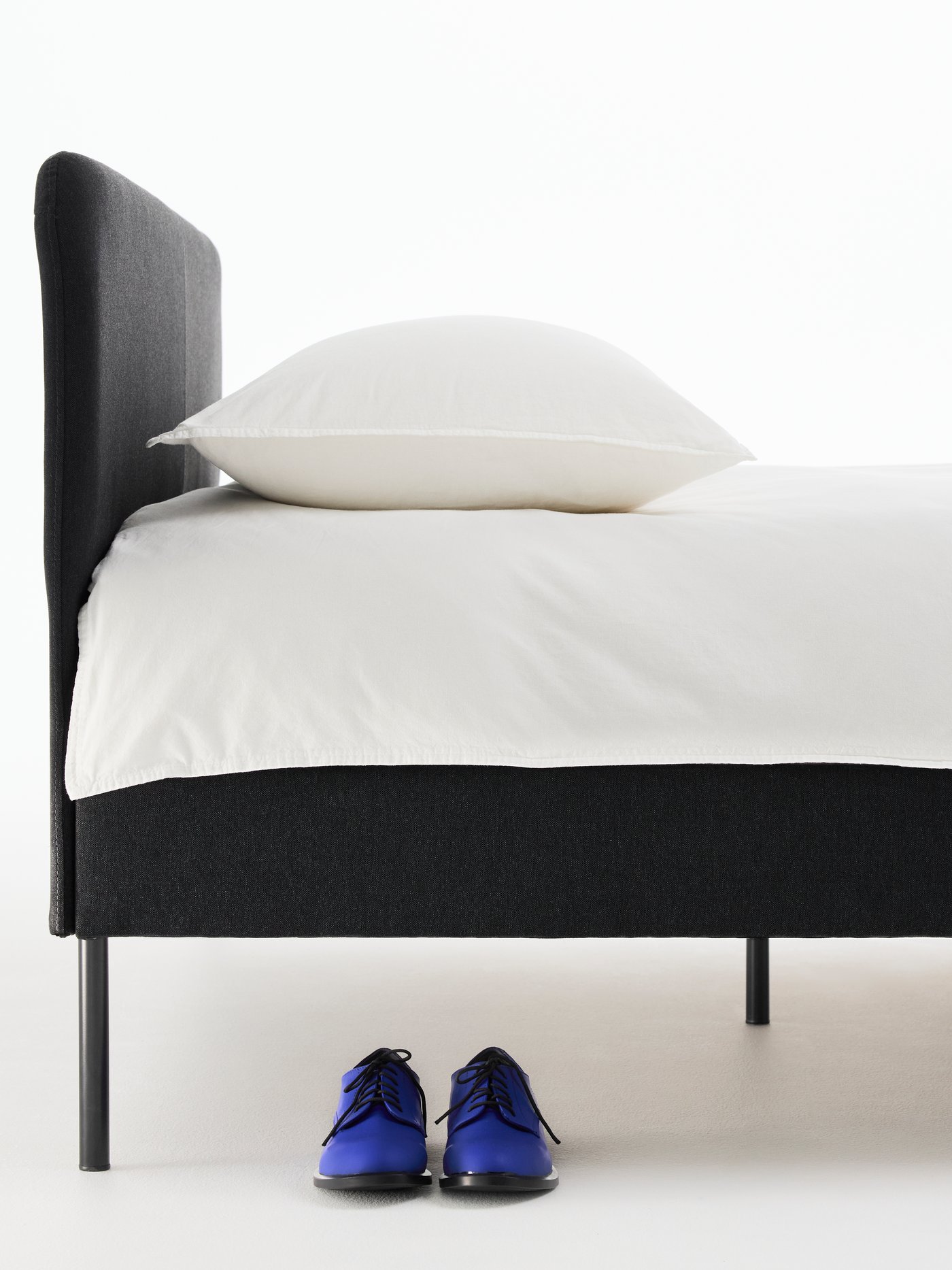Close-up of a black SLATTUM bed, neatly made with white bed linen. A pair of blue dress shoes is underneath the bed.