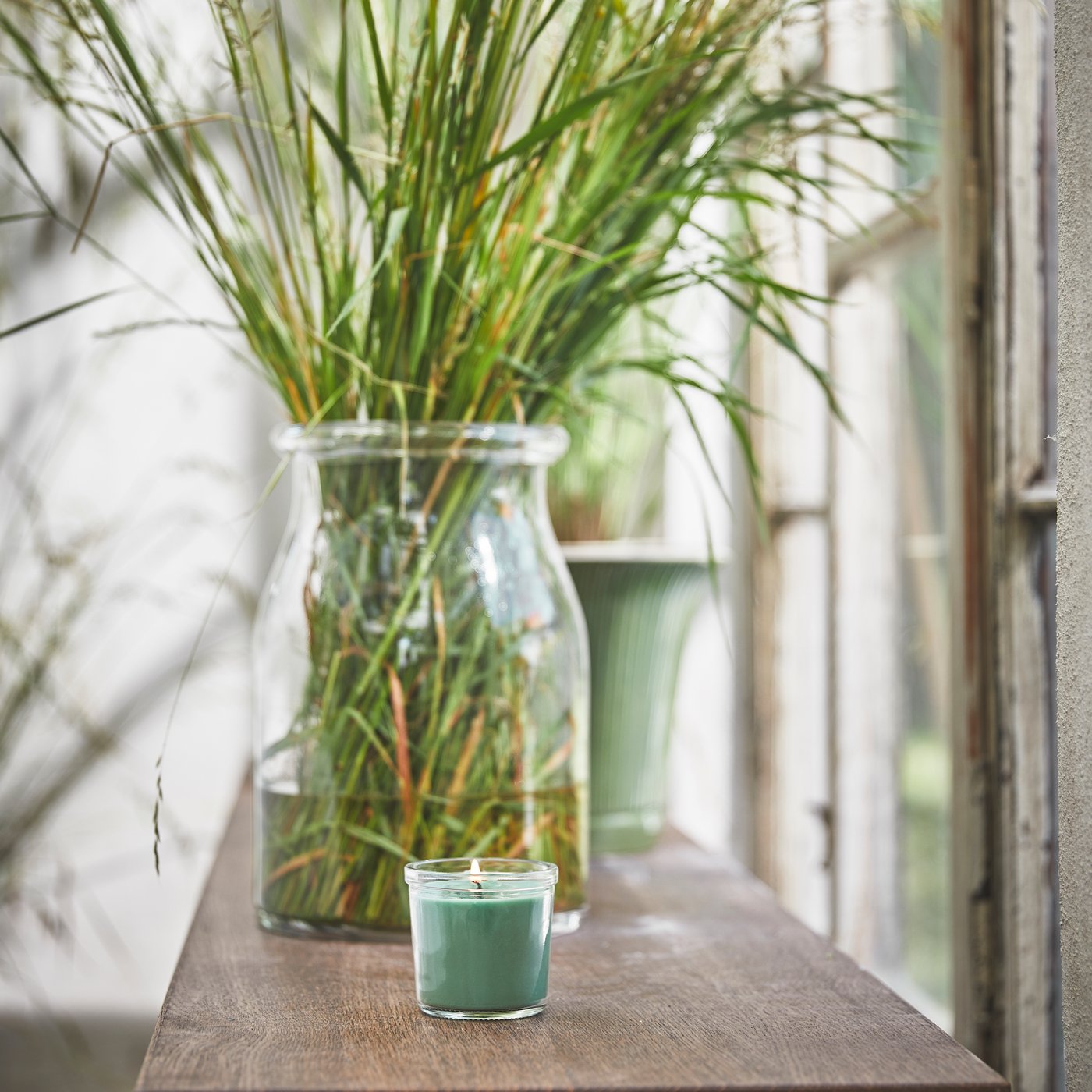 A light grey HEDERSAM scented candle with the scent of fresh grass. On the background, there's a BEGÄRLIG vase with high grass.